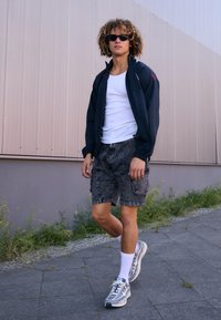 Black lightweight jacket, white tank top, gray patterned shorts, white socks, and gray sneakers. Curly hair and sunglasses, walking on pavement.