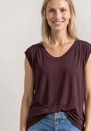 Femme souriante aux cheveux blonds lisses, portant un haut sans manches violet foncé et un jean bleu sur un fond uni.