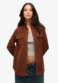 Bruine corduroy shirt met twee borstzakken, knoopsluiting en een stoffen label. Draag het over een grijze tanktop en hoge denim.