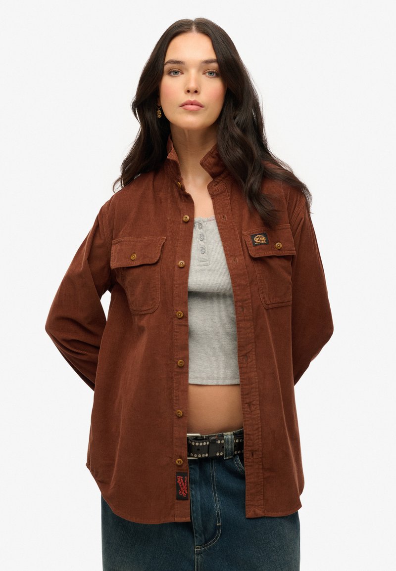 Bruine corduroy shirt met twee borstzakken, knoopsluiting en een stoffen label. Draag het over een grijze tanktop en hoge denim.