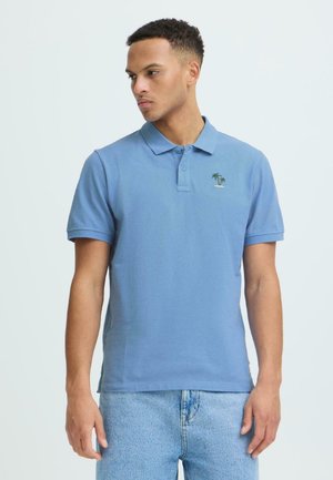 Junger Mann trägt ein hellblaues Poloshirt mit kleiner Palme-Stickerei, kombiniert mit hellblauen Jeans, schaut nach links vor einem schlichten Hintergrund.
