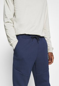Pantalones de chándal azul marino con tejido texturizado, que cuentan con una cintura con cordón y bolsillos laterales. Combinados con una camiseta de manga larga en un tono beige claro.