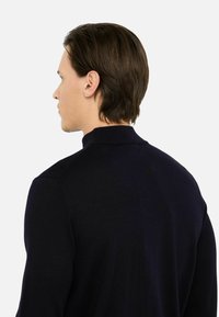Maglione blu navy a maglia con colletto alto, polsi a coste e texture liscia, mostrato da dietro su uno sfondo neutro.