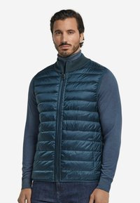 Mörkblå pufferväst med horisontell söm, hög krage och framzipper. Bärs över en långärmad blå skjorta med fickor.