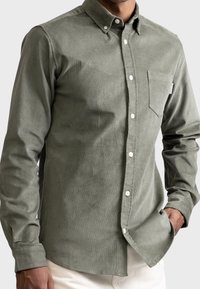 Camicia in corduroy verde oliva con colletto button-down, taschino frontale e bottoni bianchi. Presenta una superficie texture e maniche lunghe.