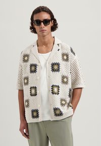 Gehaakte shirt in beige met geometrische patronen in marineblauw, grijs en olijfgroen. Heeft een knoopsluiting aan de voorkant en korte mouwen. Lichtgewicht stof.
