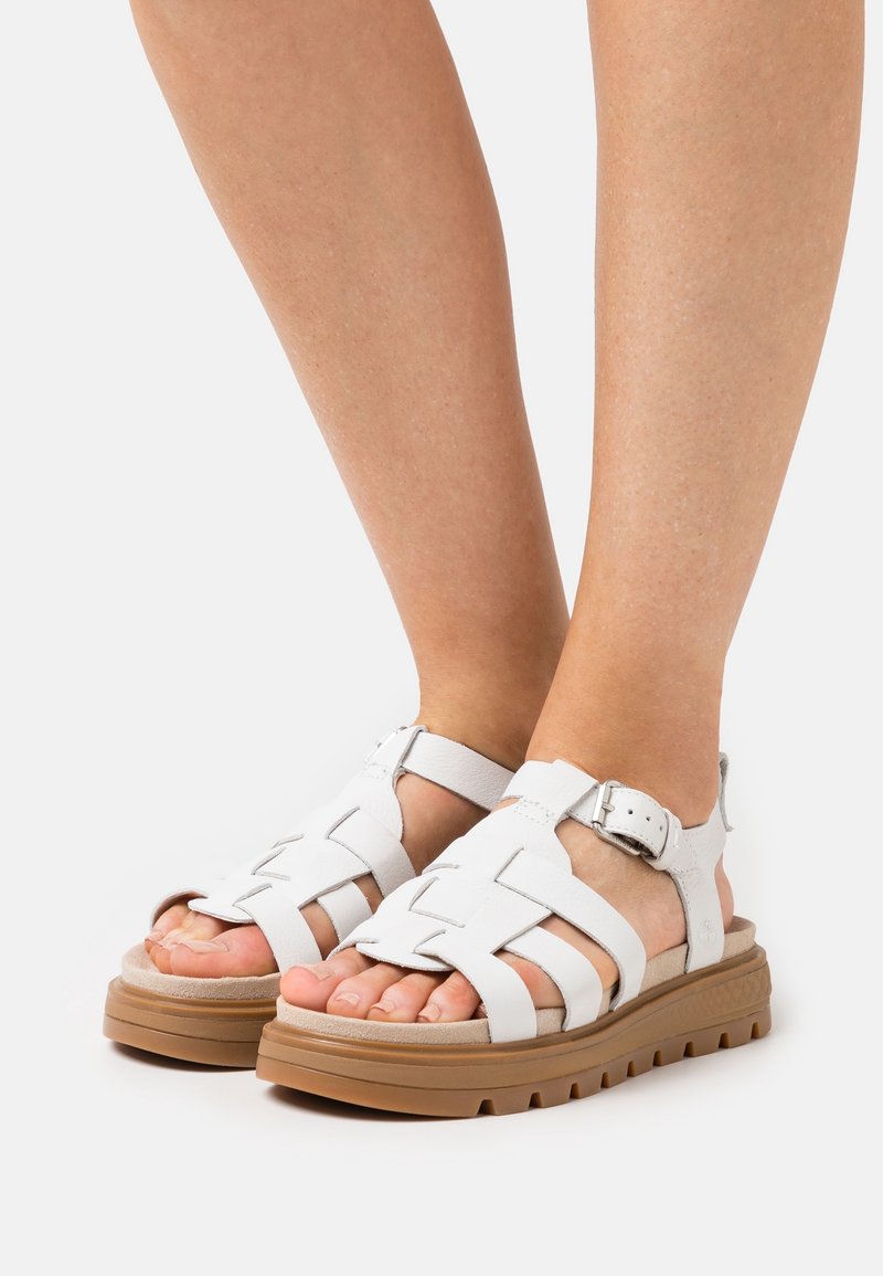 Pieds portant des sandales en cuir blanc avec des semelles épaisses couleur tan et des brides à la cheville, posés sur une surface blanche.