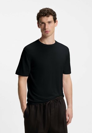 Mann mit kurzen lockigen Haaren steht frontal, trägt ein schlichtes schwarzes T-Shirt mit kurzen Ärmeln und weite dunkle Hosen vor weißem Hintergrund.
