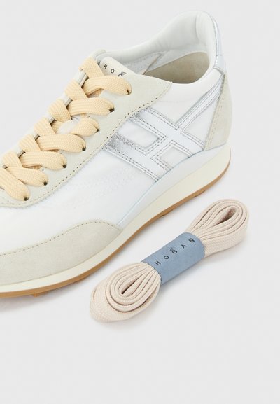 Λευκό και μπεζ αθλητικό παπούτσι με μπεζ κορδόνια, σουέτ λεπτομέρειες, ασημένιο λογότυπο στο πλάι και ένα επιπλέον ζευγάρι μπεζ κορδόνια δίπλα του σε λευκό φόντο.