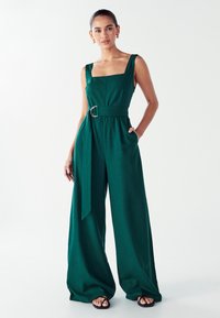 Mujer vestida con un mono sin mangas de color verde oscuro, con cinturón y pantalones de pierna ancha, usando sandalias negras, de pie contra un fondo blanco liso.