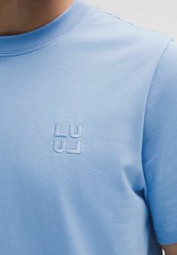 Vaaleansininen puuvillainen t-paita, jossa pyöreä aukko ja lyhyet hihat. Rintakehän alueella hienovarainen, kohokuvioitu logo. Sileä pinta.