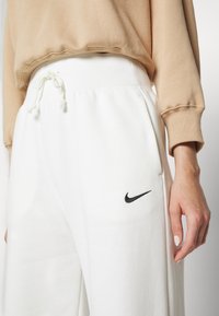 Hvite Nike joggebukser med ribbestrikket liv, snor og sidelommer. Har en svart Nike-logo på venstre lår og en myk tekstur.