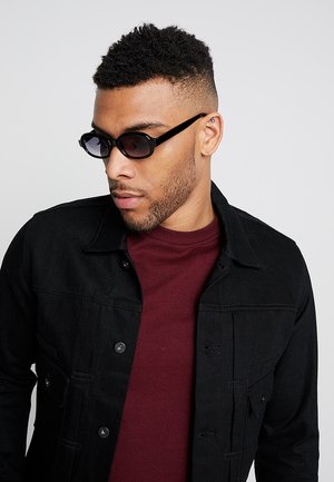 Veste en denim noir avec fermeture à boutons et poches poitrine, associée à un t-shirt col rond bordeaux. Lunettes de soleil noires au design angulaire.