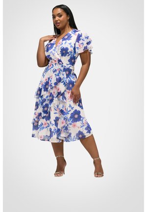 Femme portant une robe portefeuille blanche avec des imprimés floraux bleus et roses, des manches flottantes et des volants superposés, associée à des escarpins dorés à bride cheville.