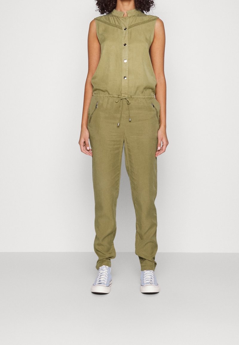 Combinaison sans manches vert olive avec boutons sur le devant, taille à cordon, poches zippées, portée avec des baskets claires sur fond blanc.