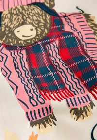 Farebný textil s medveďom v ružovom svetri a námorníckym modrým a červeným károvaným šálom. Detaily obsahujú strapce a cik-cak vzory.