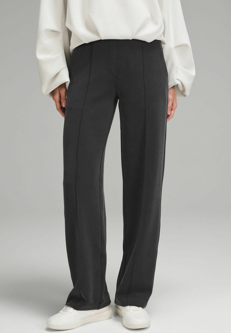 Personne portant un pantalon noir droit, un chemisier blanc à manches longues et des baskets blanches, debout sur un fond gris uni.