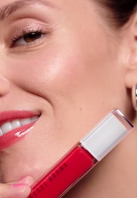 Rossetto liquido rosso in un tubo trasparente con un tappo bianco. La modella ha la pelle liscia e un colore di labbra rosa lucido, che evidenzia il rossetto.