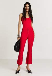 Tuta fitted rosso con scollo rotondo, gambe svasate e cerniera frontale. Gli accessori includono una borsa nera e bracciali metallici.