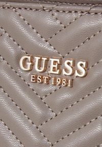 Gray quilted leather material with gold-tone "GUESS EST 1981" logo. Features detailed stitching and a smooth, glossy texture.