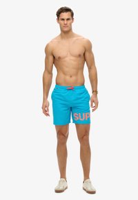 Bañadores turquesa con gráfico rosa "SUP", cintura elástica con cordón ajustable y bolsillos laterales. El material parece ligero y transpirable.