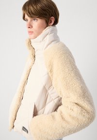 Jacket puffer bej cu mânecă din blană texturată și pufoasă. Prezintă un guler înalt, închidere cu fermoar și buzunare laterale.