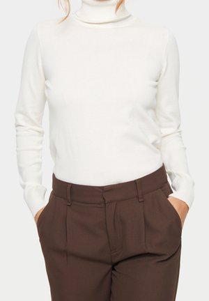 Frau trägt einen weißen, gerippten Rollkragenpullover, der in eine hochtaillierte, braune Faltenhose gesteckt ist, mit den Händen in den Taschen.