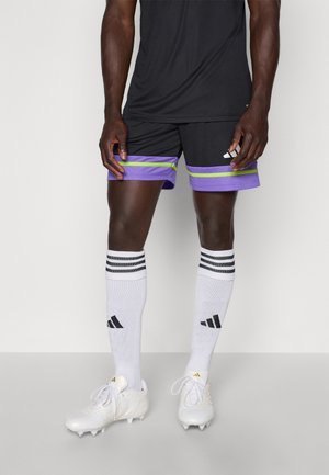 Mężczyzna ubrany w czarne i fioletowe sportowe szorty, białe, sięgające kolan skarpetki Adidas oraz białe korki, stoi na jednolitym tle.