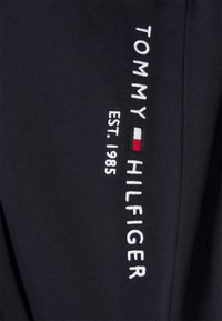 Svart tyg med vit broderad text: "TOMMY HILFIGER EST. 1985" med en färgad logodetalj i rött, vitt och blått.