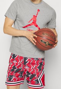 Camiseta de algodón gris con un logo rojo de "Jumpman". Shorts de baloncesto con patrón rojo y negro y rayas blancas. Sosteniendo un balón de baloncesto Spalding.