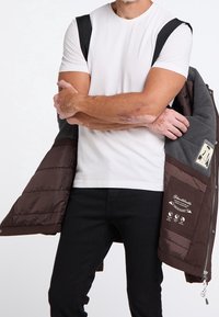 Brown puffer jacket with gray accents, featuring zip closure and interior branding details, worn over a white t-shirt and black jeans.
