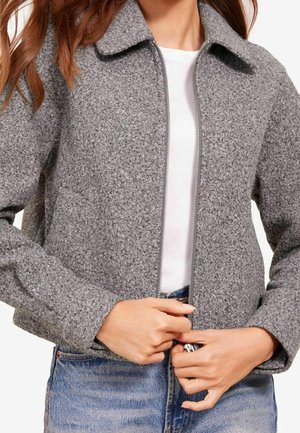 Femme fermant une veste en laine grise texturée par-dessus une chemise blanche, portant un jean bleu.