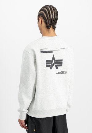 Person with braided hair wears light gray sweatshirt featuring black Alpha Industries logo and text on the back, paired with black pants.