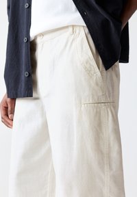 Beige pants with textured fabric, featuring side pockets and a piped accent. Worn with a black, button-down shirt.