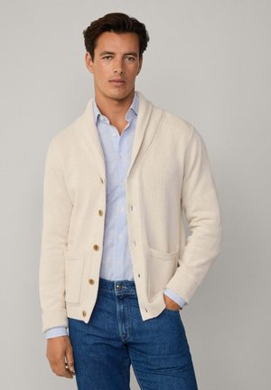 Hellbeige Strickjacke mit Schalkragen, Knopfverschluss und zwei vorderen Taschen. Trägt man über einem blau-weißen gestreiften Hemd, kombiniert mit Jeans.