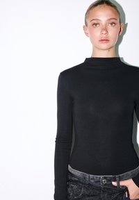 Musta pitkähihaine paita korkealla kauluksella, valmistettu sileästä kankaasta, yhdistettynä teksturoituihin tummiin farkkuihin, joissa on yksinkertainen napitus.