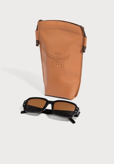 Pochette en cuir marron avec logo MCM embossé à côté de lunettes de soleil rectangulaires noires avec verres bruns sur fond blanc.