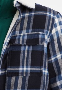 Camicia di flanella con motivo a quadri blu e navy con dettagli bianchi, colletto bianco morbido e due tasche sul petto. Base layer verde visibile sotto.