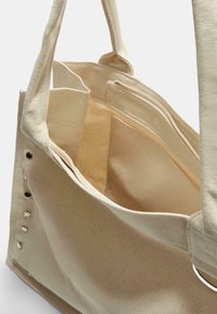 Beige kangaskassi, jossa on kaksi avointa taskua ja vetoketjullinen lokero, varustettu metallisilla yksityiskohdilla ja teksturoidulla pinnalla.