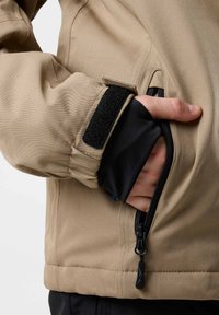 Beige Jacke mit einer Reißverschlusstasche und einem Klettverschluss am Ärmel. Sie zeichnet sich durch eine glatte Textur und ein kontrastierendes schwarzes Innenfutter aus, das funktionale Details präsentiert.