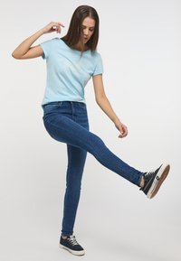 Jasnoniebieska bawełniana koszulka z graficznym nadrukiem, połączona z ciemnoniebieskimi dopasowanymi dżinsami i czarnymi trampkami, prezentowana w dynamicznej pozie.