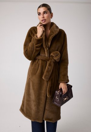 Une femme porte un long manteau de fourrure marron ceinturé, en tenant son col et en portant un sac à main foncé sur un fond de studio uni.