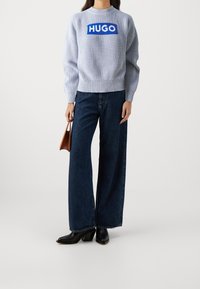 Vrouw in een lichtblauwe gebreide trui met het "HUGO" logo, donkere wijde jeans, zwarte laarzen en een kleine bruine handtas.