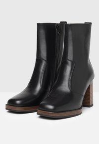 Stivali alla caviglia in pelle nera con una texture liscia, chiusura con zip laterale e punta quadrata. Presentano un tacco in legno rigato marrone.