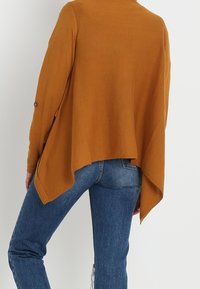 Langdrogige, mosterdkleurige cardigan met een gedrapeerde achterkant. Zachte textuur, knoopdetail op de mouwen, gecombineerd met blauwe spijkerbroeken.