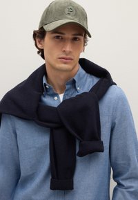 Maglione a maglia blu marinato legato intorno al collo sopra una camicia celeste con bottoni; cappello di velluto a coste verde con una lettera 'P' ricamata.