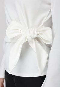 Άσπρο ύφασμα δεμένο σε φιόγκο στη μέση ενός λευκού μακρυμάνικου τοπ, φορεμένο με μαύρο παντελόνι.