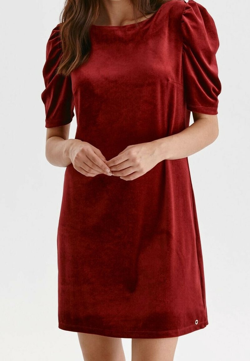 Femme portant une robe en velours rouge à manches courtes avec des épaules bouffantes, debout les mains jointes librement devant elle.