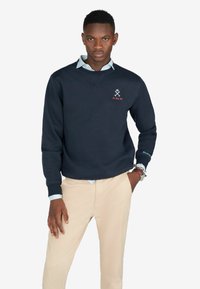 Sudadera azul marino con cuello redondo y puños acanalados. Presenta un pequeño logo bordado en rojo y blanco en el pecho. Lleva puestos pantalones beige.