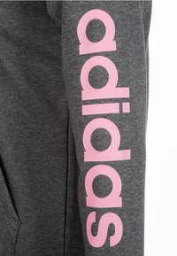 Tissu de sweat-shirt gris à texture douce, avec un lettrage rose vif "adidas" imprimé verticalement le long de la manche gauche.